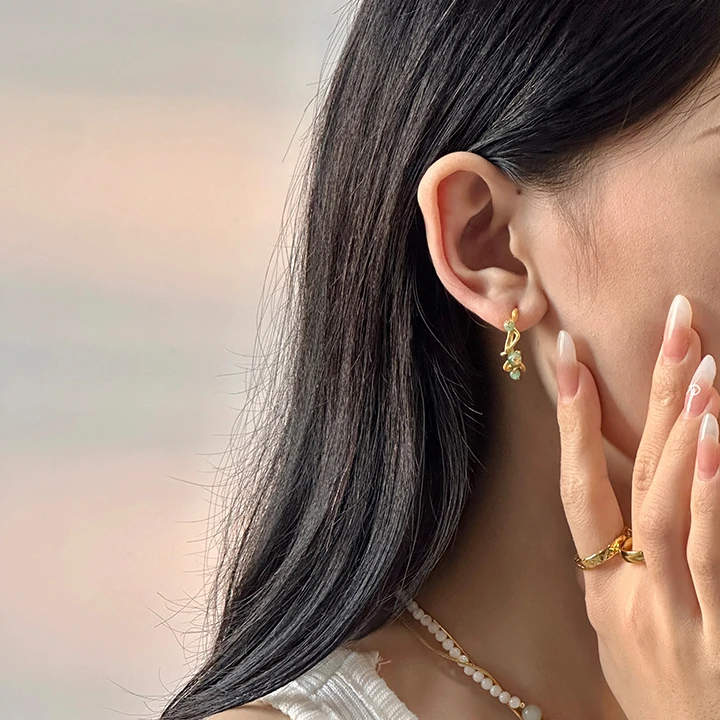 Woman wearing LIVKE gold vine earrings with green gemstones at outdoor garden party, showcasing organic luxury jewelry
